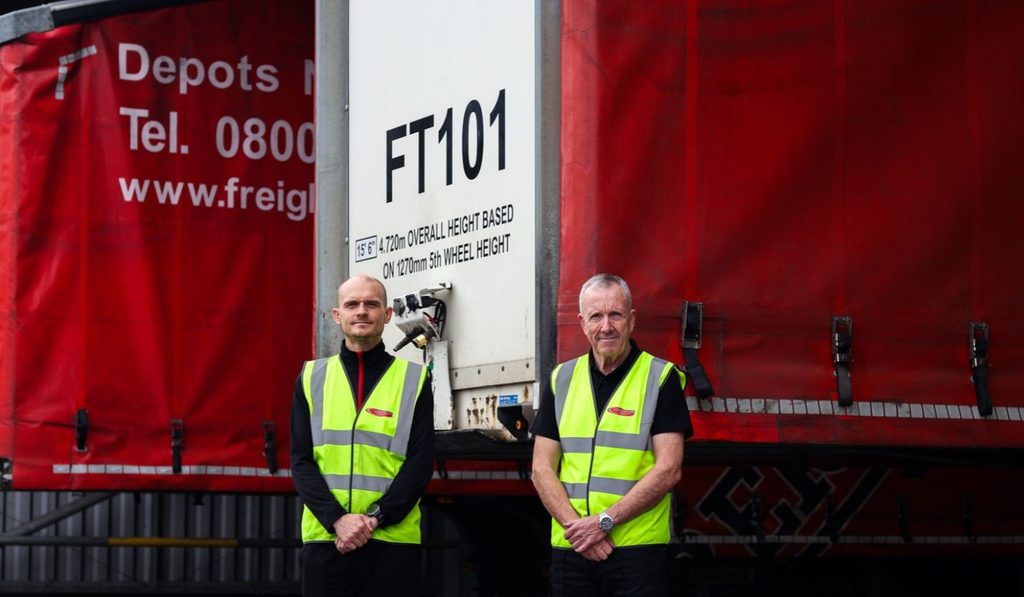 2 men with custom branded hi-vis vests standing in front of 2 lorries
